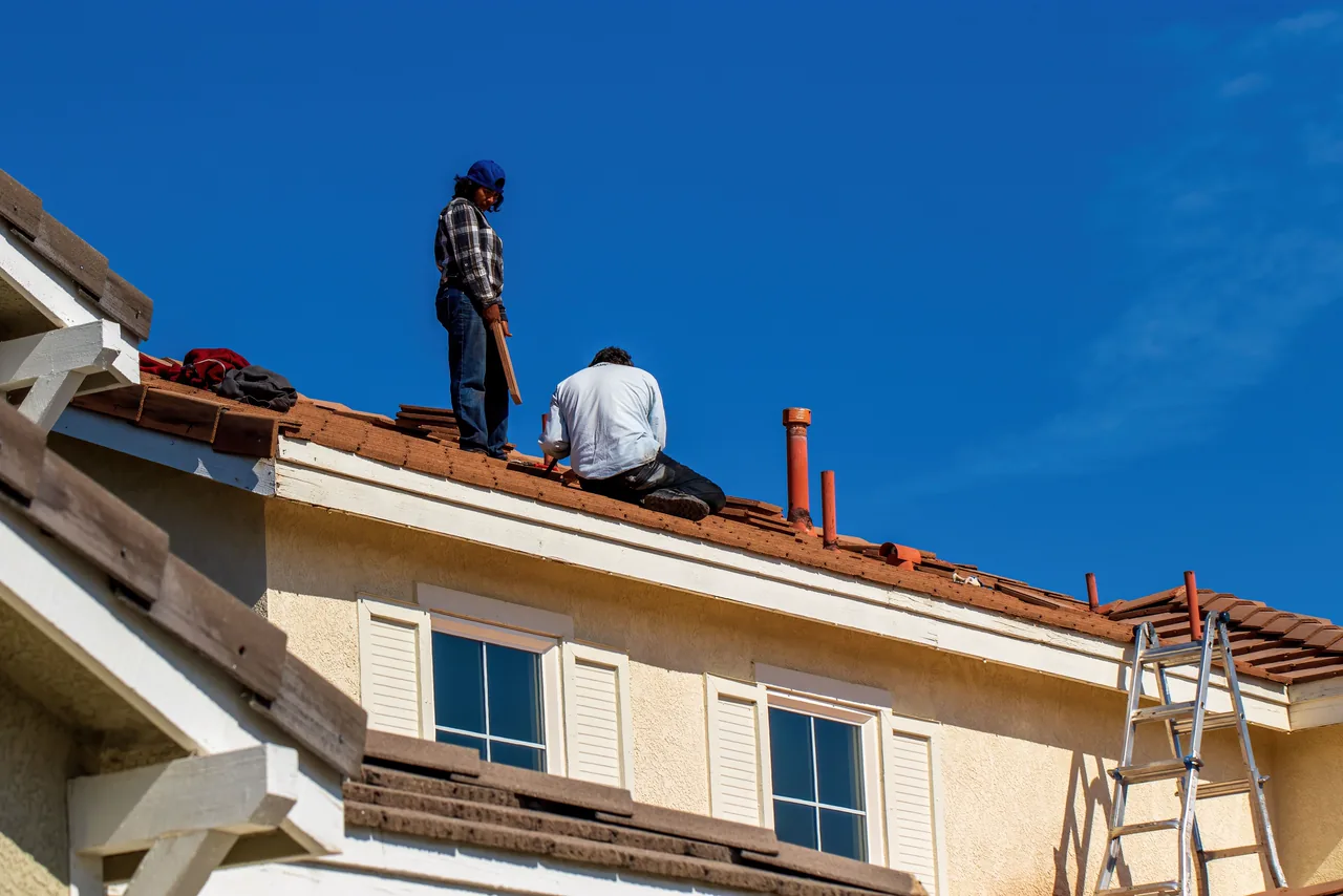 Two roofers laying clay tiles on the roof of a two-storey stucco home